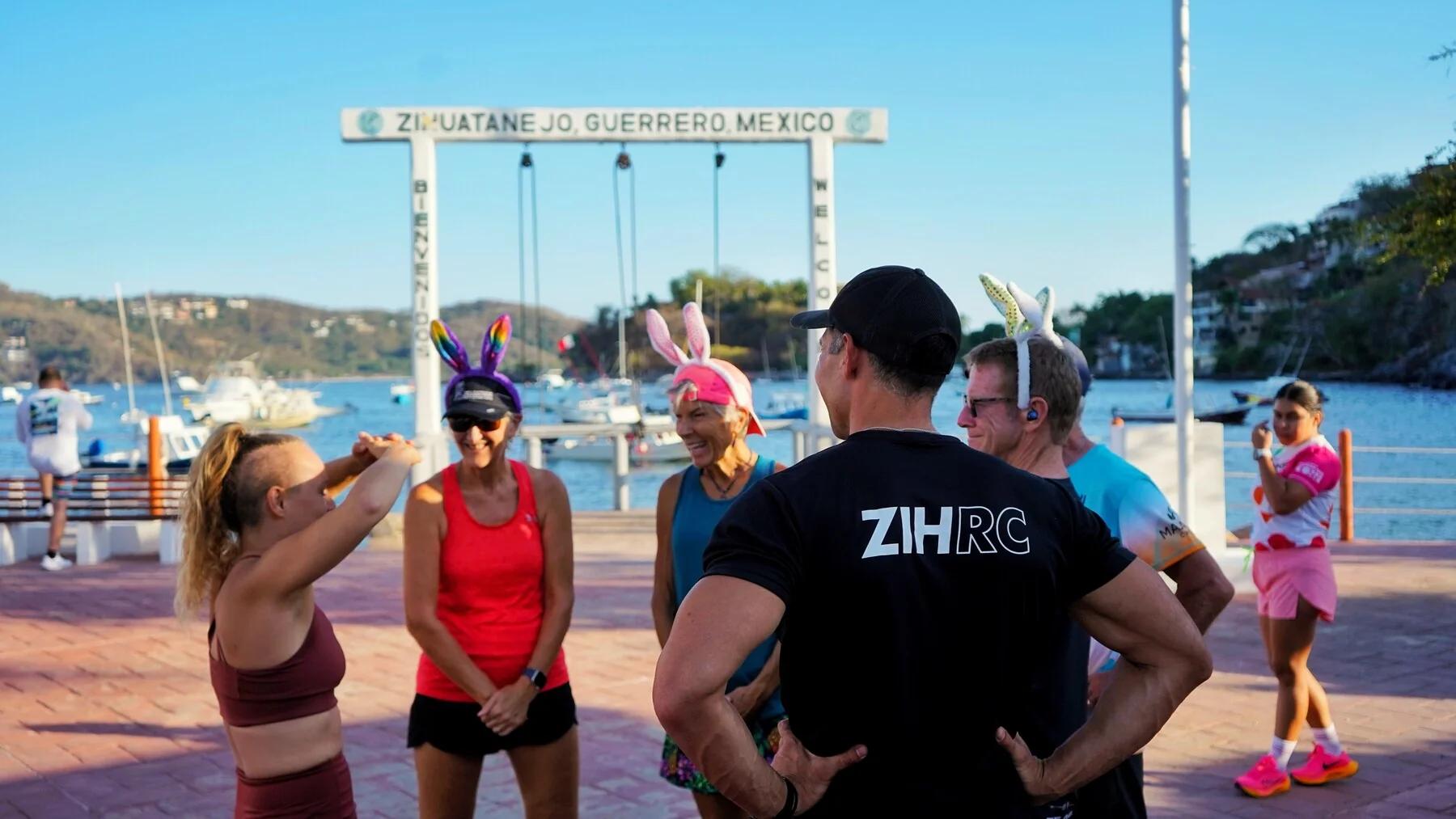 Corredores del Zihuatanejo Run Club conversando bajo el arco de bienvenida a Zihuatanejo, Guerrero, con la bahía y los barcos al fondo